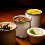 Three enamel mugs with assorted soups garnished attractively on a warm wood table.