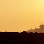A couple embraces on a rocky shore with ships in the background during a stunning sunset.