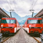 Two red trains at a Swiss station with mountains in the background.