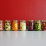 A row of vibrant, multicolored mason jars filled with assorted fermented vegetables against a red wall.