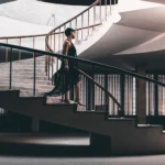 An elegant woman in a black dress gracefully walking up a spiral staircase indoors.