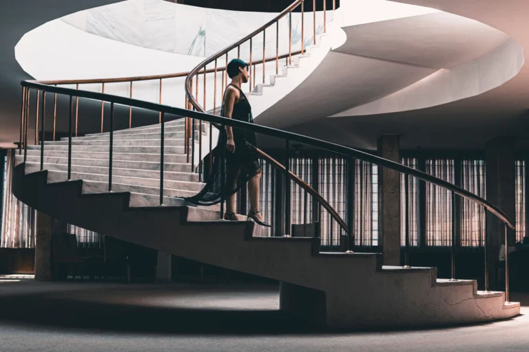 An elegant woman in a black dress gracefully walking up a spiral staircase indoors.
