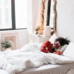 Woman wearing a sleep mask, resting on a cozy white bed surrounded by indoor plants in a bright bedroom.
