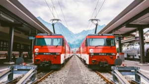 Two red trains at a Swiss station with mountains in the background.