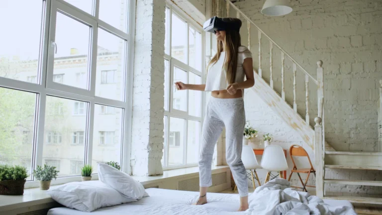 Young woman exploring virtual reality while standing on bed in a modern, sunlit room.