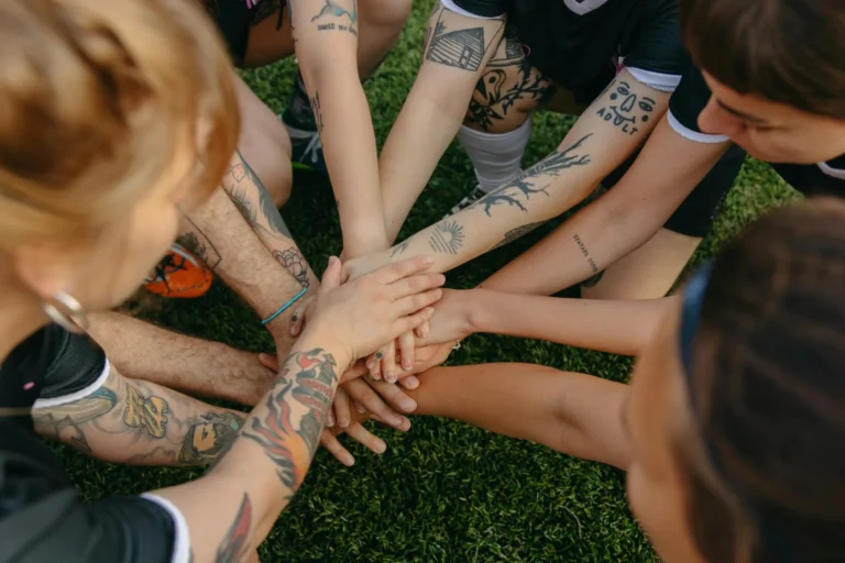 Tattooed team members join hands in a circle signifying unity and teamwork on a grassy field.