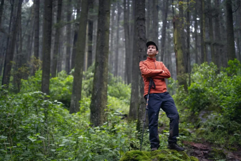 A hiker stands confidently in a lush forest, arms crossed, portraying exploration.