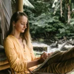 Woman in hammock sketching in a peaceful outdoor setting by a river.