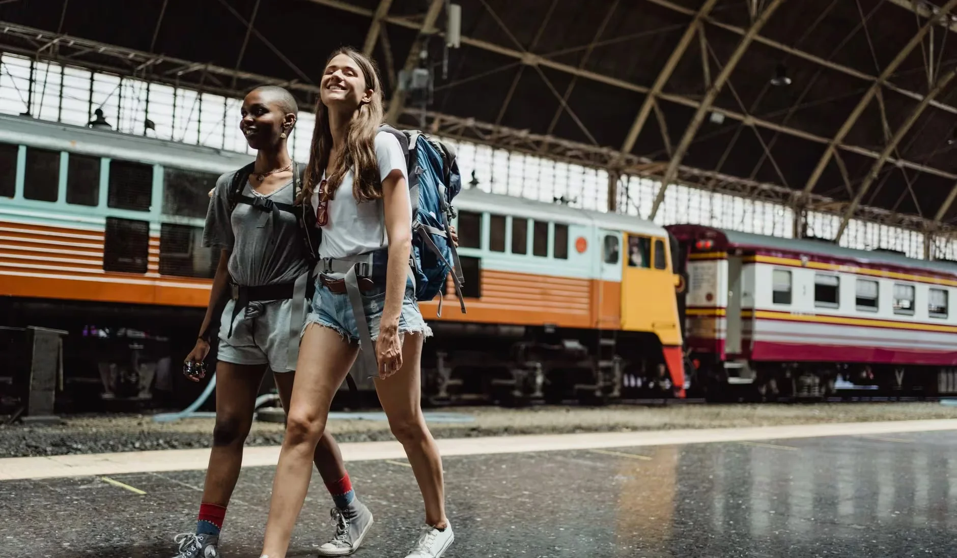 Two women with backpacks walking and smiling at a railway station.