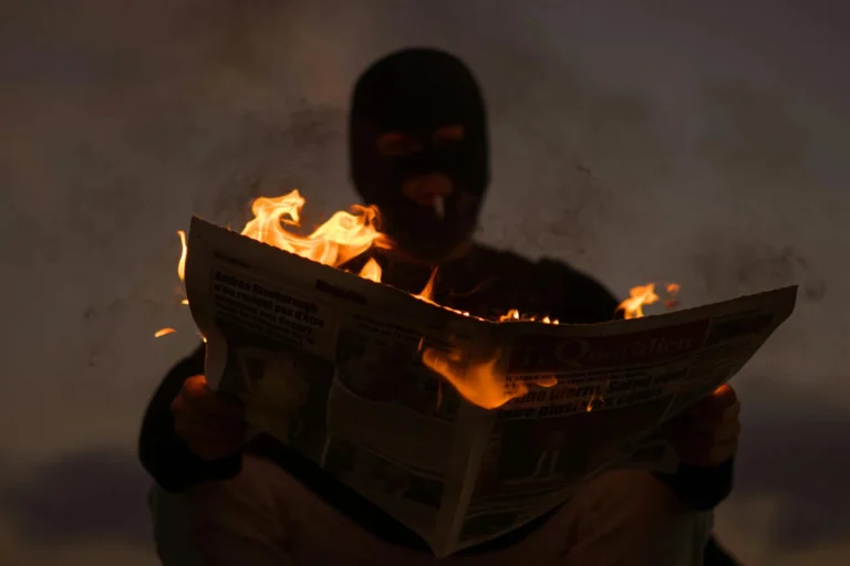 A masked person holds a burning newspaper in dramatic low light, evoking mystery.