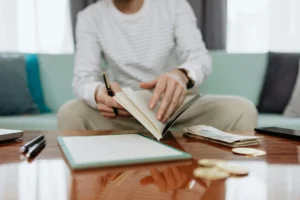 Individual reviewing finances at a table with notebook, money, and laptop.