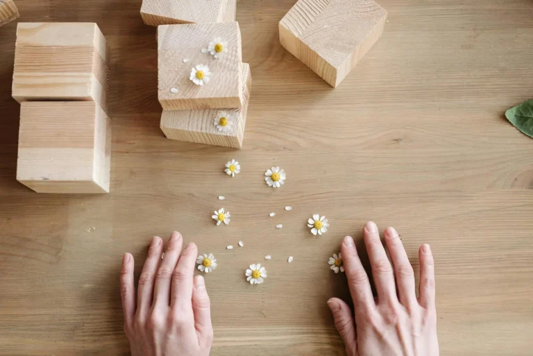 Hands surrounded by wooden blocks and chamomile flowers on a wooden surface.