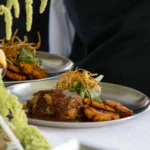 A delicious gourmet dish with meat, fried plantains, and greens, served on a silver platter.