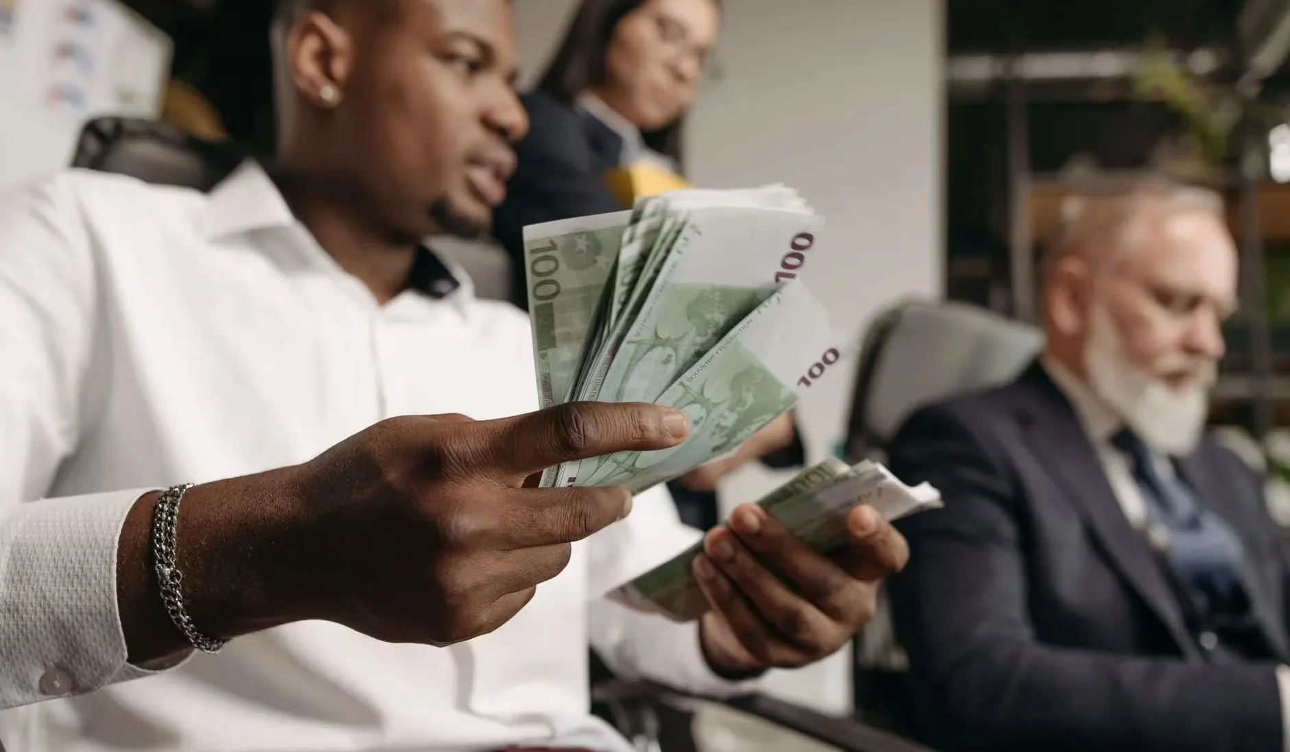Three professionals in business attire counting cash in a modern office setting.