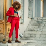 Stylish young woman in red suit standing by an ornate wooden door in an urban setting.