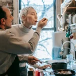 Side view of content ethnic man in casual clothes and apron feeding happy wife while preparing delicious dinner at stove in kitchen