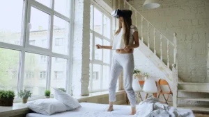 Young woman exploring virtual reality while standing on bed in a modern, sunlit room.