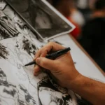 Artist drawing detailed comic book characters with a pen at a lively event.