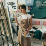 A female artist in a studio, pondering her artwork with a brush and canvas.