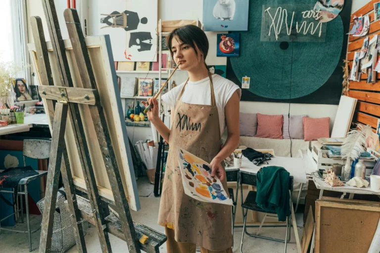 A female artist in a studio, pondering her artwork with a brush and canvas.