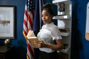 Professional woman holding a book poses confidently in an office setting with flags in the background.