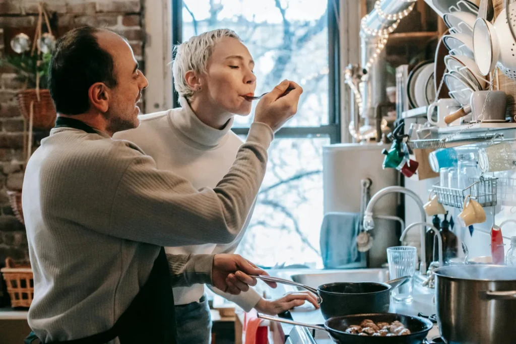 Side view of content ethnic man in casual clothes and apron feeding happy wife while preparing delicious dinner at stove in kitchen