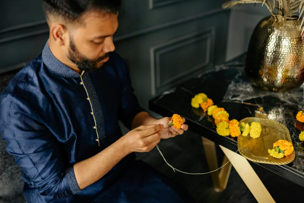 Man in traditional attire making a marigold garland indoors, focusing on craft.