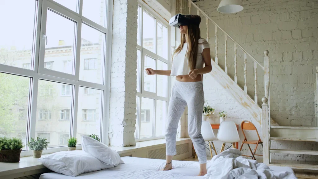 Young woman exploring virtual reality while standing on bed in a modern, sunlit room.