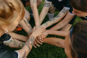 Tattooed team members join hands in a circle signifying unity and teamwork on a grassy field.