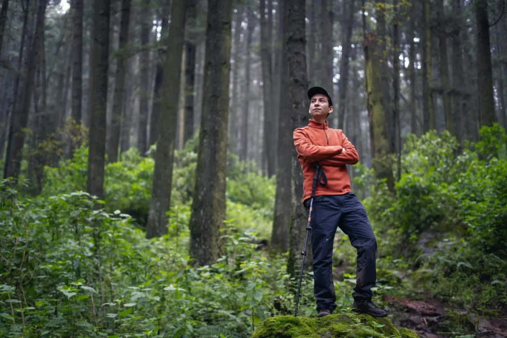 A hiker stands confidently in a lush forest, arms crossed, portraying exploration.