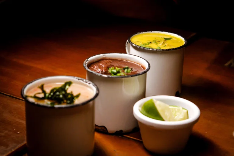 Three enamel mugs with assorted soups garnished attractively on a warm wood table.