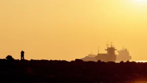 A couple embraces on a rocky shore with ships in the background during a stunning sunset.