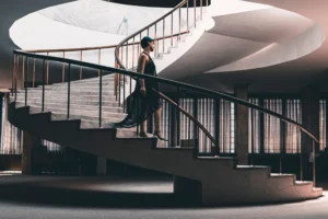 An elegant woman in a black dress gracefully walking up a spiral staircase indoors.