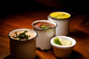 Three enamel mugs with assorted soups garnished attractively on a warm wood table.