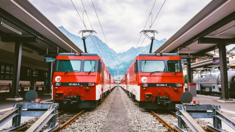 Two red trains at a Swiss station with mountains in the background.