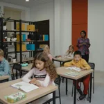 Elementary students focus on classwork with a teacher overseeing in a bright classroom.