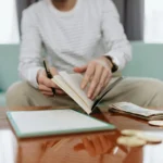 Individual reviewing finances at a table with notebook, money, and laptop.