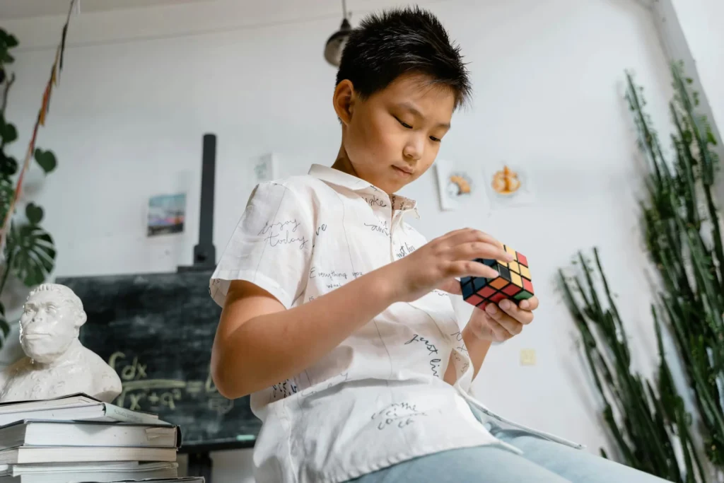 Asian boy thoughtfully solving a Rubik's Cube in a classroom, surrounded by books and a classroom setting.