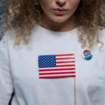 A woman with curly hair holding an American flag and wearing a vote pin against a blue background.