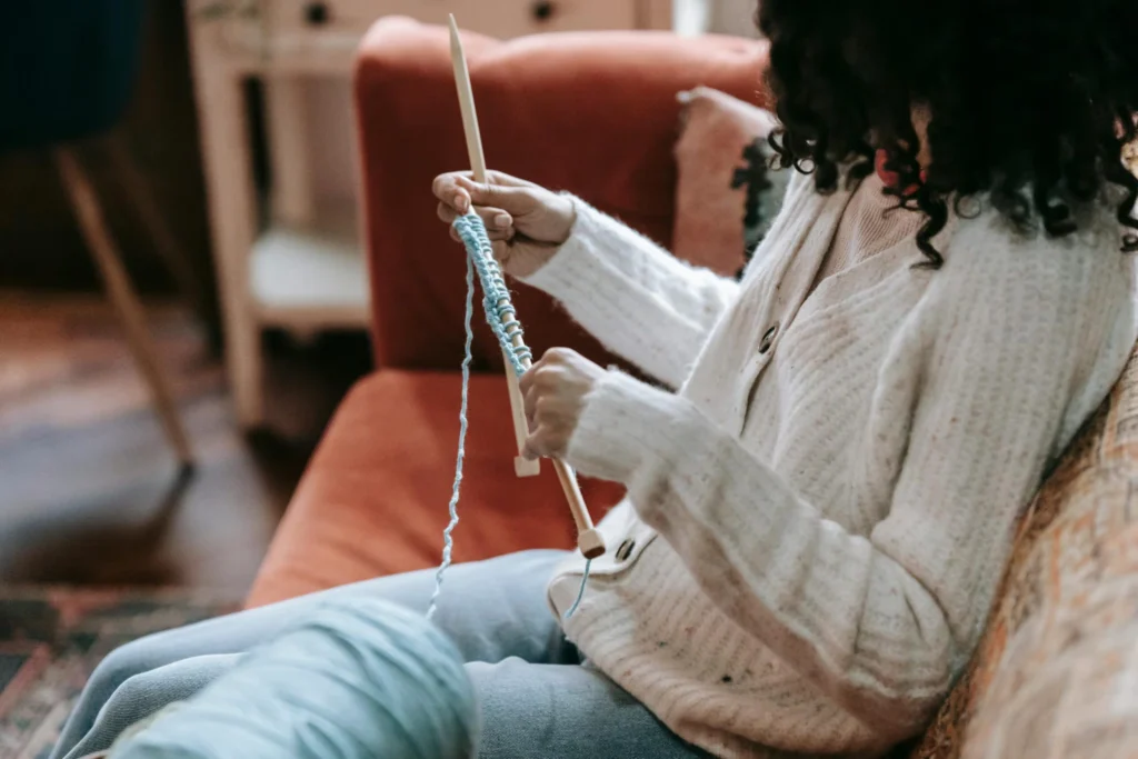 Side view of crop anonymous female artisan with yarn and needles knitting on couch in living room