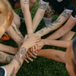 Tattooed team members join hands in a circle signifying unity and teamwork on a grassy field.