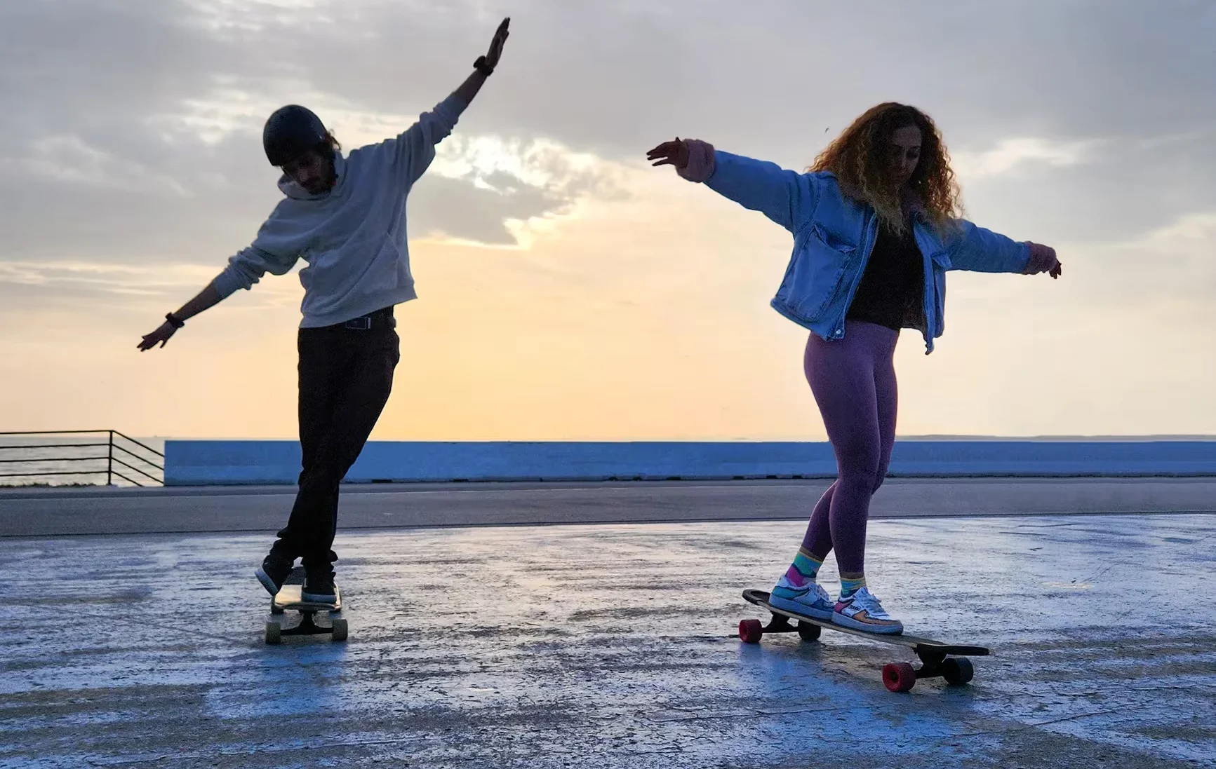 Two young adults skateboarding gracefully on a seaside promenade at sunset in İstanbul, Türkiye.