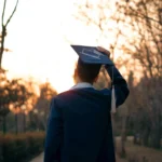 A graduate in a cap and gown celebrates outdoors during sunset.