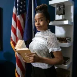 Professional woman holding a book poses confidently in an office setting with flags in the background.