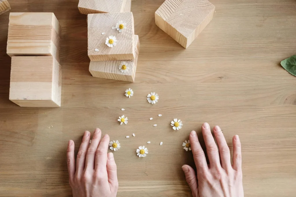 Hands surrounded by wooden blocks and chamomile flowers on a wooden surface.