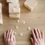Hands surrounded by wooden blocks and chamomile flowers on a wooden surface.
