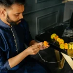 Man in traditional attire making a marigold garland indoors, focusing on craft.