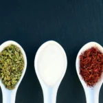 Three ceramic spoons with herbs, salt, and saffron on dark slate background.