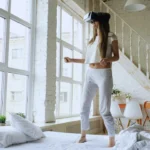 Young woman exploring virtual reality while standing on bed in a modern, sunlit room.