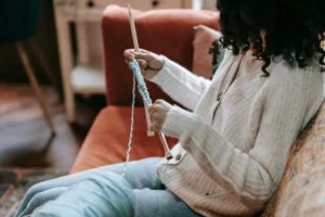 Side view of crop anonymous female artisan with yarn and needles knitting on couch in living room